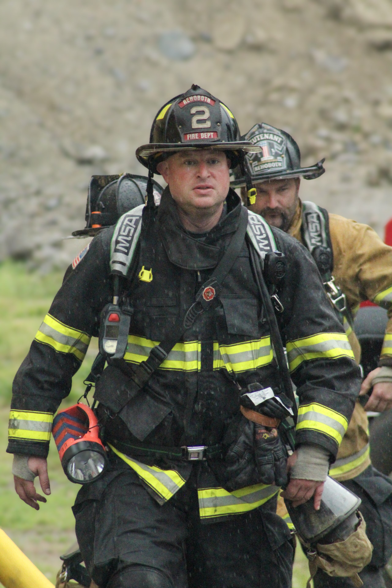 Bryan Syrett in full firefighter turnout gear and helmet outdoors during training, with rocky terrain in the background.