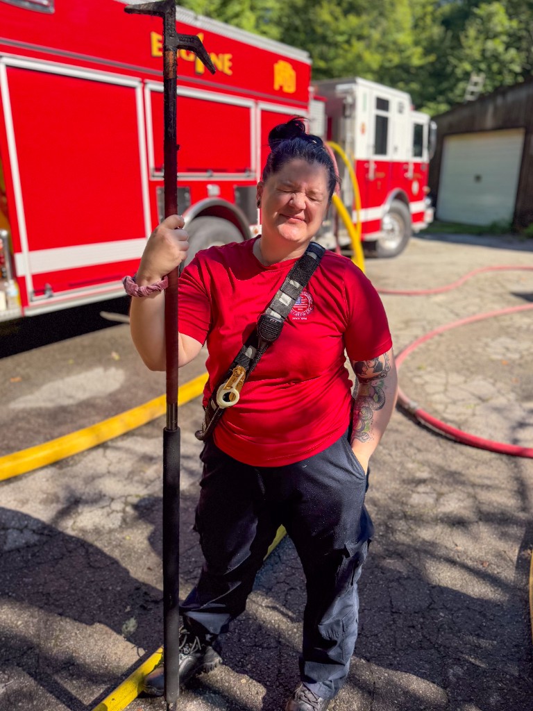 Lindsey Burnett standing in front of Engine 1 in a red department shirt, radio strap, and boots, holding a roof hook with fire hose on the ground around her.