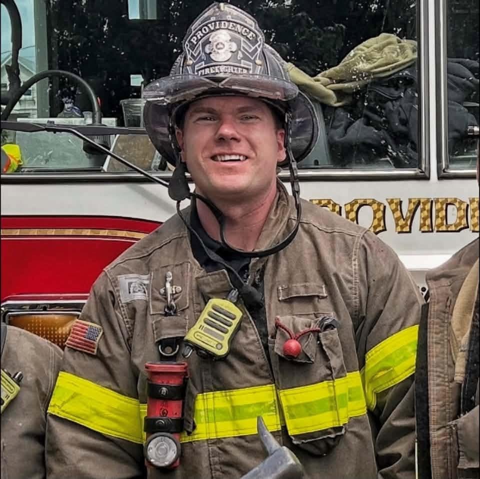 Stuart “Stu” Blakely smiling in full turnout gear and a Providence firefighter helmet, standing in front of a Providence fire truck.